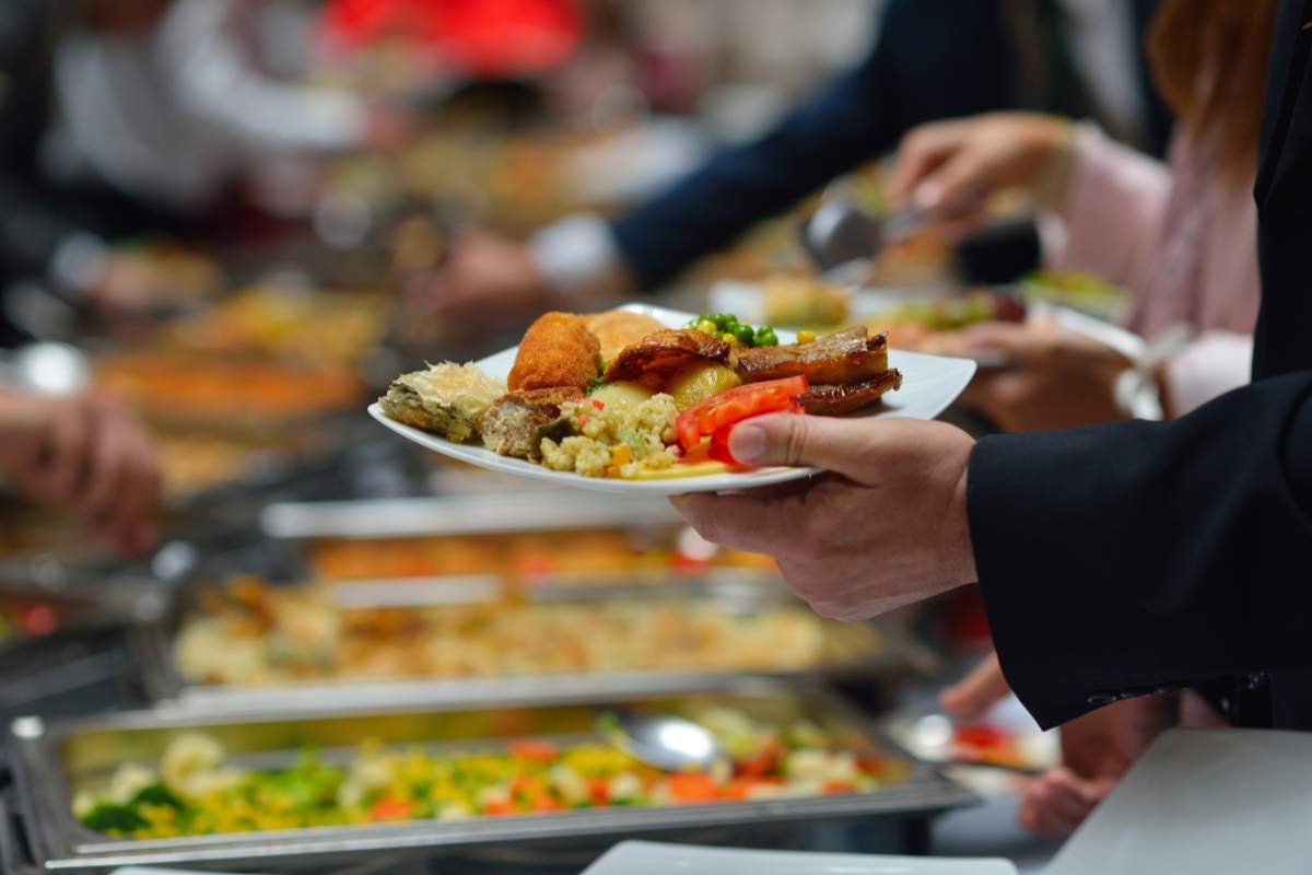 People serving themselves a variety of dishes from a buffet-style meal, courtesy of our exceptional office catering solutions, with one person holding a plate filled with various foods in the foreground.