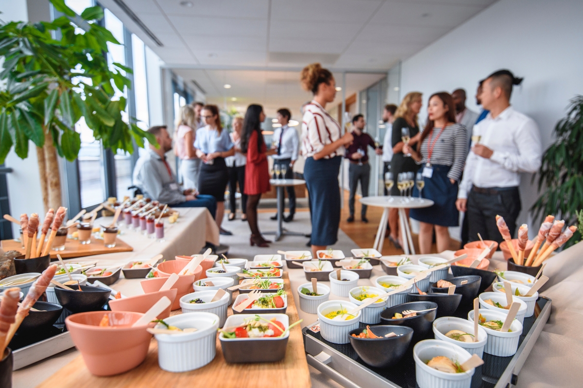 A group of people socializing and standing in a room with a long table covered with various food dishes in the foreground, showcasing office catering solutions. There are plants and large windows in the background.