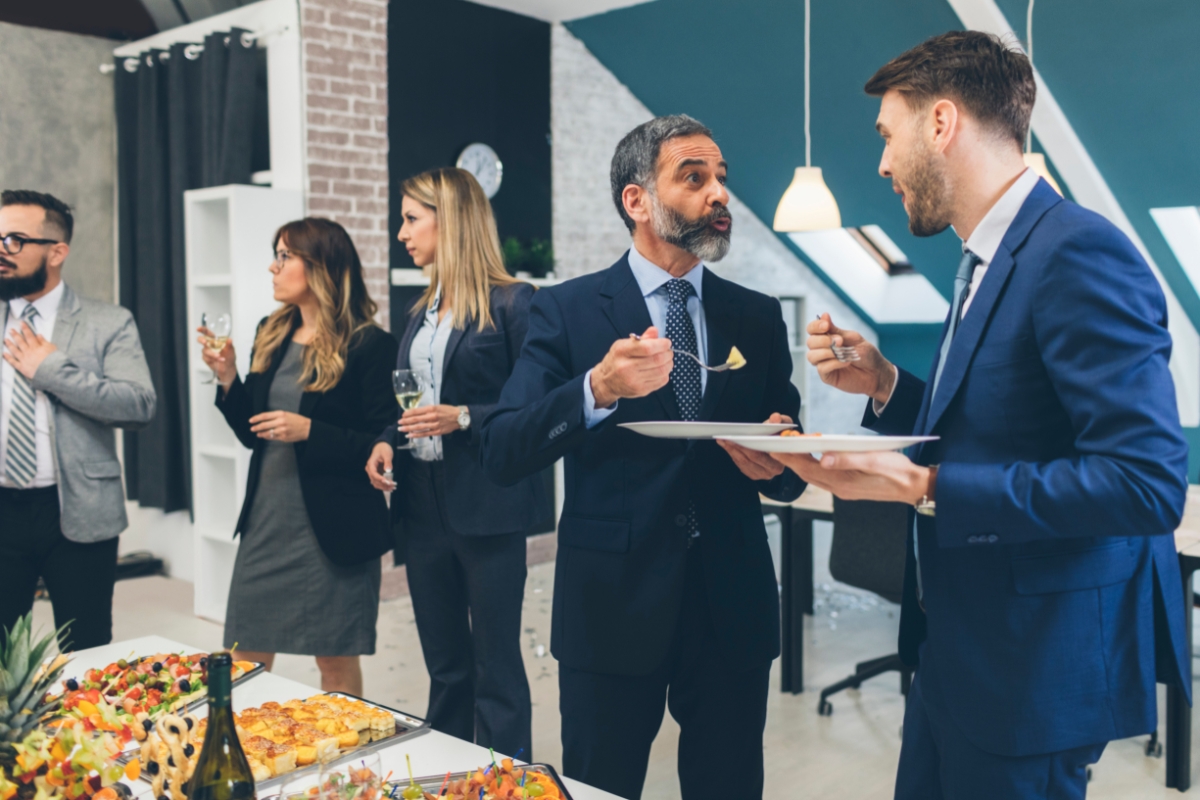 A group of professionally dressed people are standing and conversing in an office setting. They are holding plates of food and drinks provided by office catering solutions, with a table of buffet-style refreshments in the foreground.