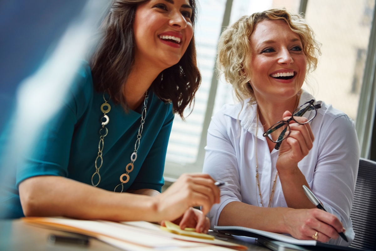 Two women sitting at a desk, smiling and engaged in conversation. One is holding eyeglasses and a pen, while the other has a notebook open in front of her. A large window is in the background, hinting at an engaging discussion on office catering solutions.