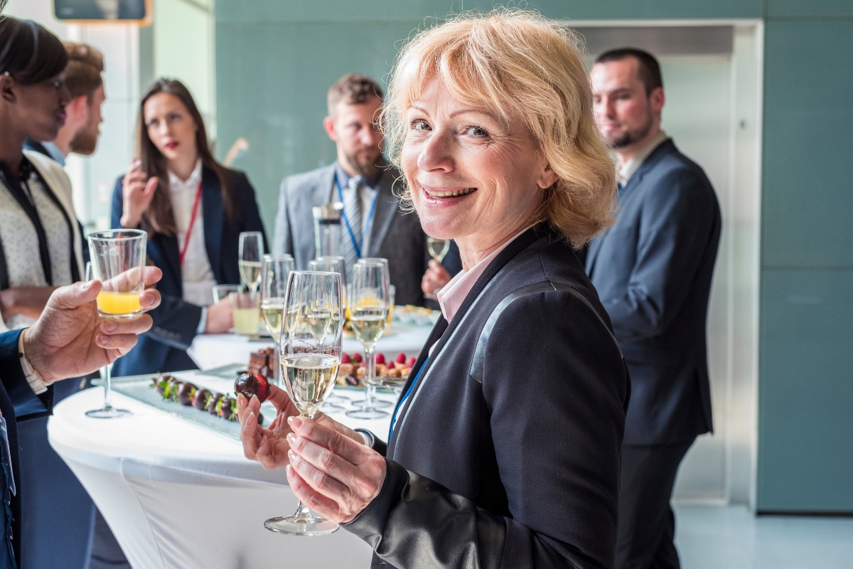 A woman in business attire holds a glass of wine and smiles at a networking event featuring office catering solutions. Other attendees are conversing in the background.