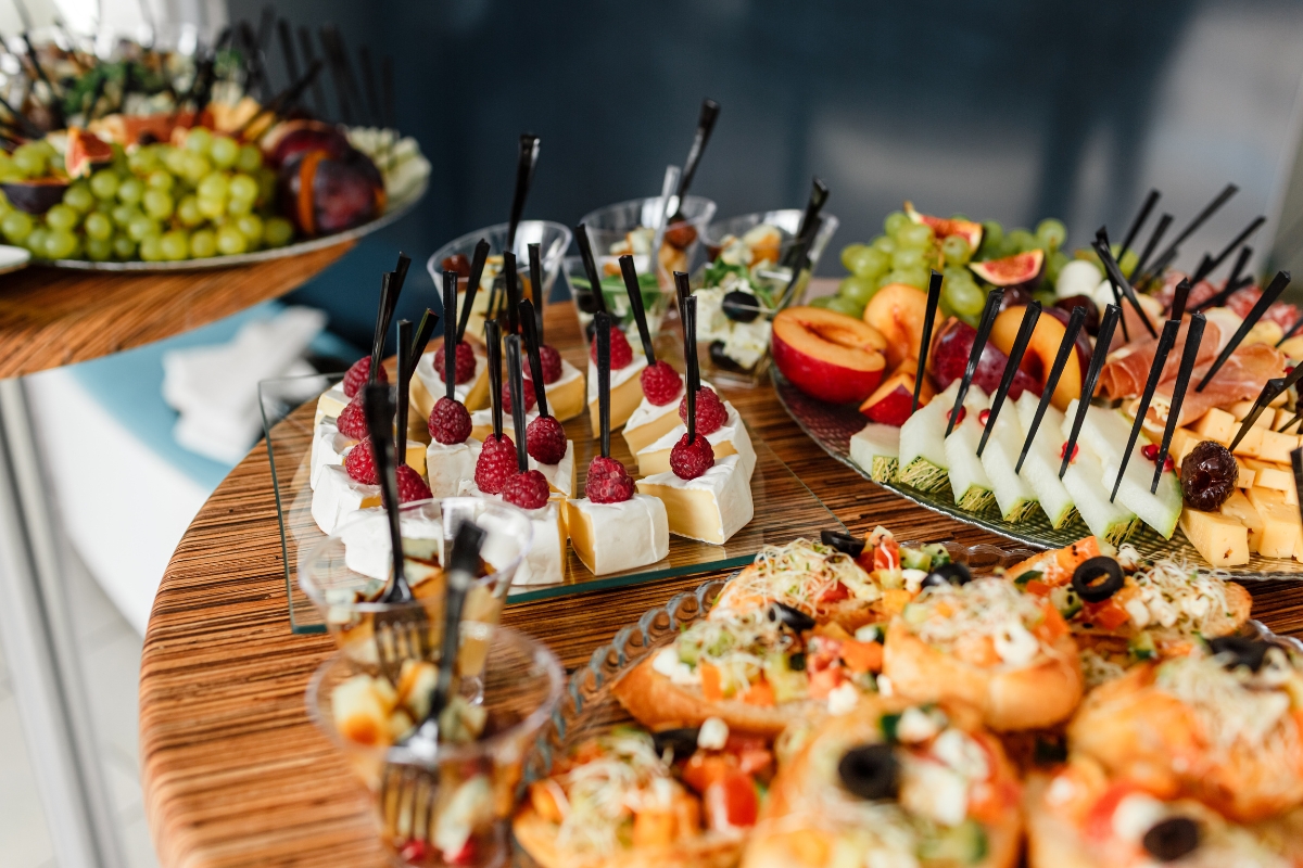 A table with an assortment of appetizers, perfect for office catering solutions, includes cheese and fruit skewers, pastry bites topped with vegetables, and a bowl of grapes and figs.