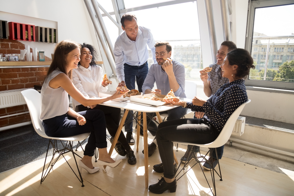 A group of six people enjoy pizza together while sitting and standing around a round table in a brightly lit room, thanks to the convenient office catering solutions.