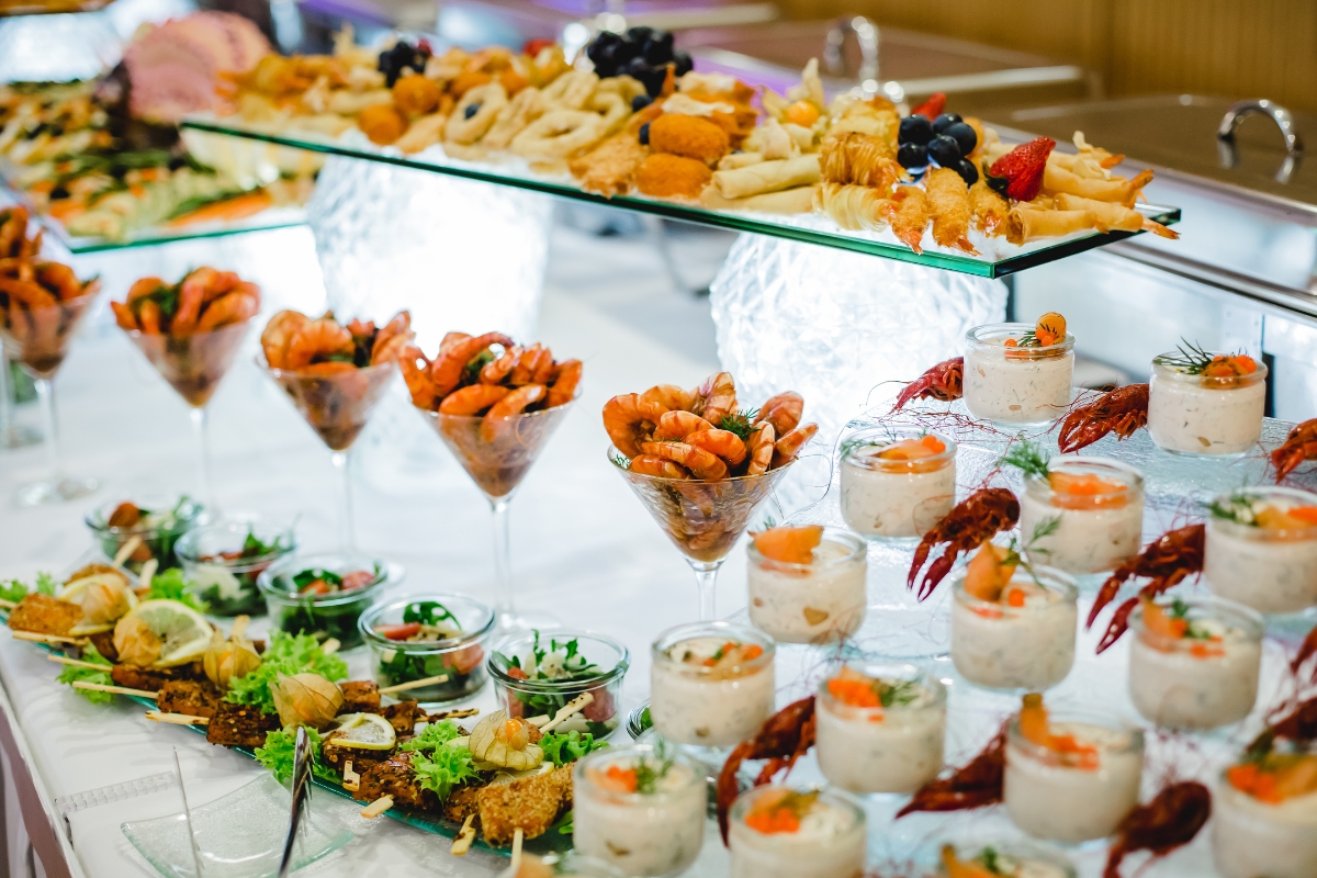 A buffet table displays an assortment of seafood appetizers in glasses and dishes, including shrimp, lobster tails, and parfaits, alongside various fried snacks and garnished platters from an eco-conscious catering service.