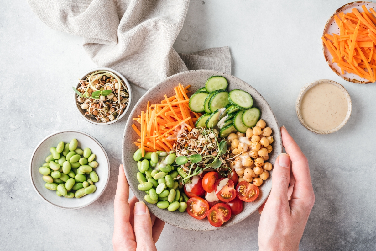 A bowl filled with sliced cucumbers, shredded carrots, cherry tomatoes, chickpeas, edamame, and sprouts. Nearby are bowls of edamame, shredded carrots, sprouts, and a beige dressing. Hands hold the bowl as part of our eco-conscious catering experience.