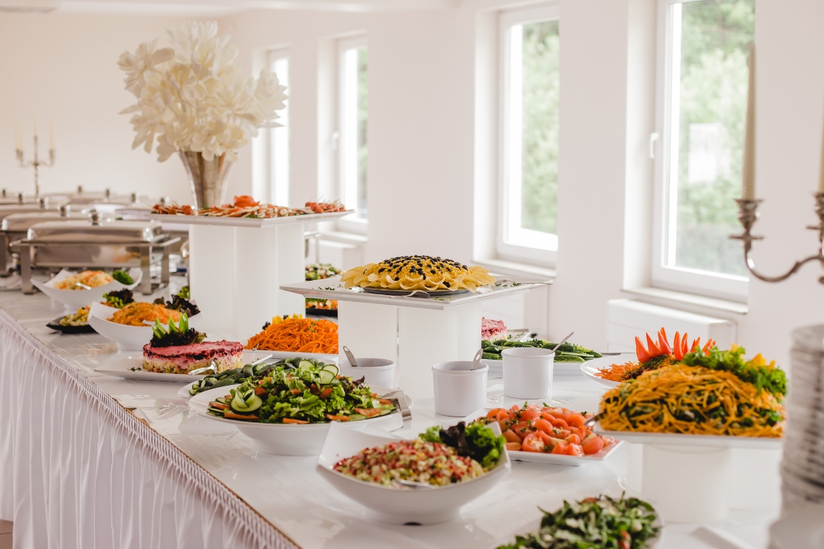 A long table is set with a variety of dishes, including salads, pasta, and garnished platters. The setup features eco-conscious catering in a bright room with large windows.