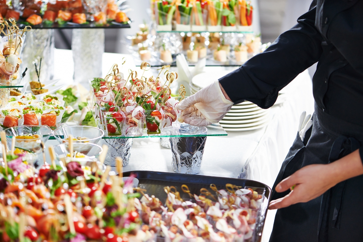 A person wearing gloves arranges appetizers and finger foods on a buffet table filled with a variety of colorful dishes, showcasing the eco-conscious catering efforts through reusable serving ware and locally sourced ingredients.