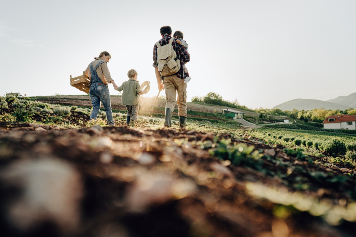 A family of four, with two adults and two children, walks through a rural farm field at sunset. The adult man carries one child on his back while the adult woman holds hands with the other child, all enjoying the serene beauty that echoes the ethos of farm to table catering.