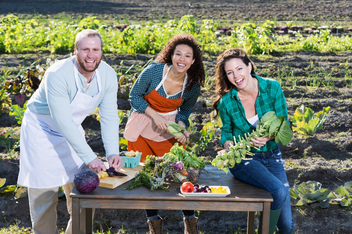 Three people cutting and arranging vegetables at a table in a garden, exemplifying farm to table catering. The man on the left is wearing a white apron, while the two women are in casual clothing. Various fresh vegetables are on the table.