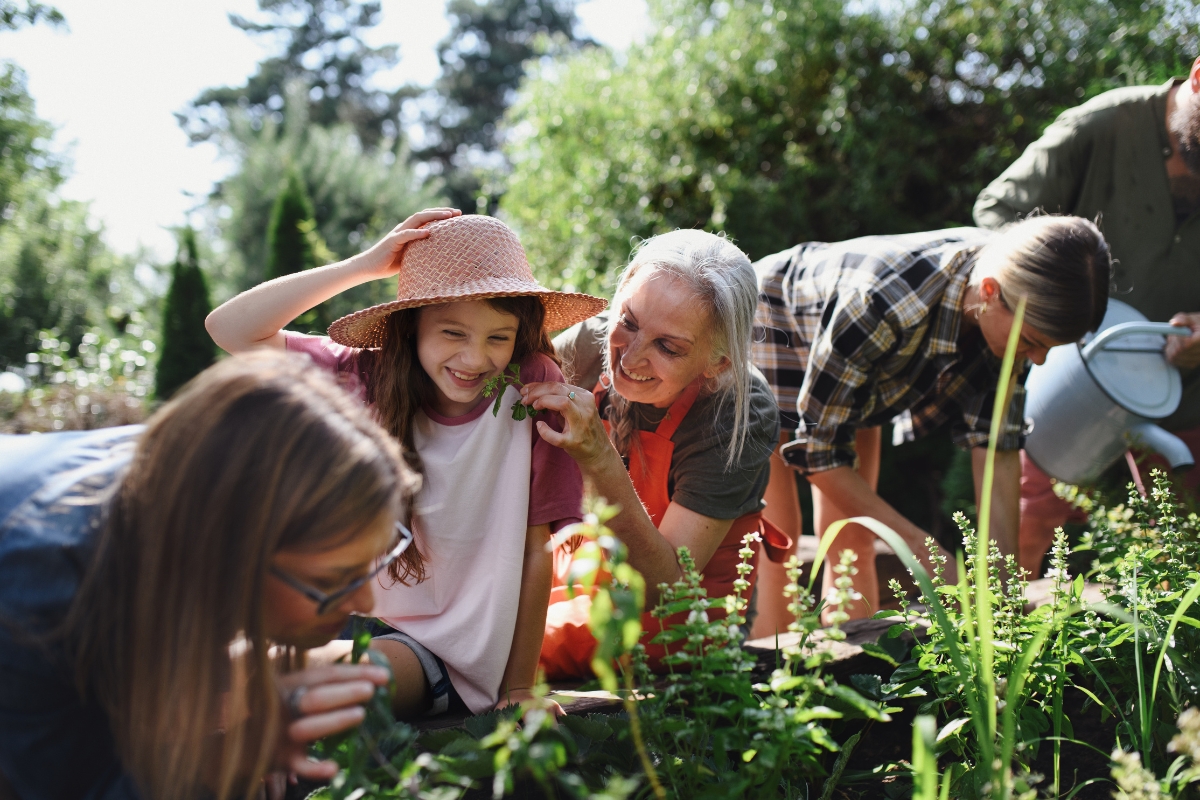 A group of people, including children and adults, are gardening together outside, surrounded by greenery. A woman with gray hair is smiling and talking with a girl wearing a hat about their farm to table catering experience.