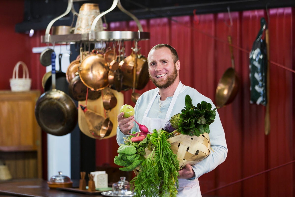 A man in a white apron stands in a kitchen holding a basket of fresh vegetables and an apple, embodying the essence of farm-to-table catering, with hanging copper pots and other kitchen items in the background.