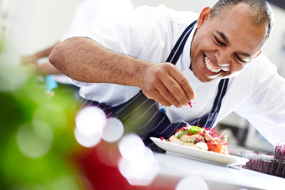 In a bright kitchen setting, a chef in a white coat and striped apron garnishes a dish with a smile, embodying the essence of custom catering.