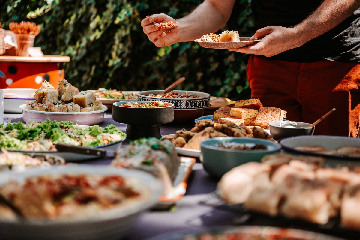 A person is serving food from a table filled with various dishes, including salads, bread, and appetizers, set outdoors—a feast brought to life through custom catering.