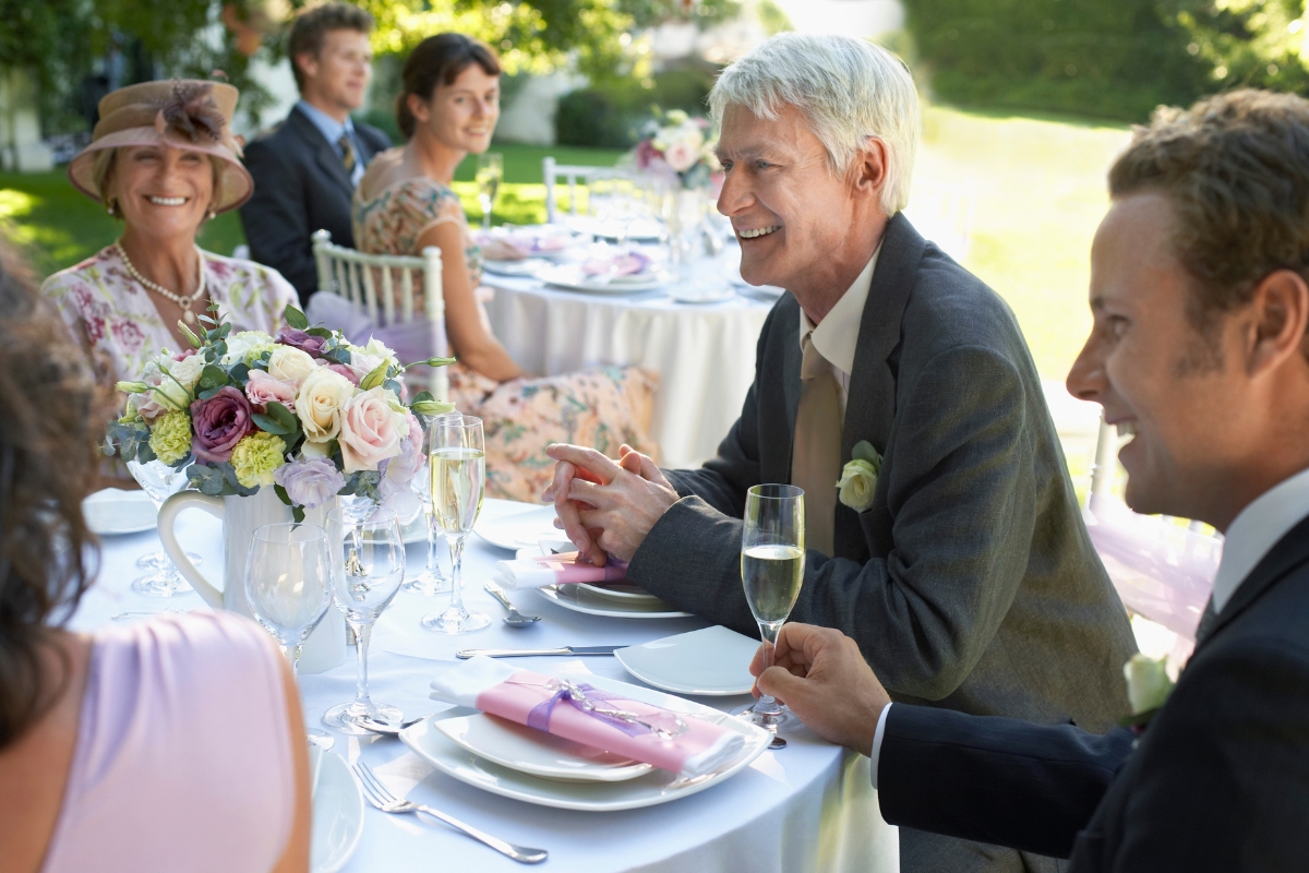 People seated at a decorated outdoor table, enjoying custom catering, dressed formally, smiling and chatting.