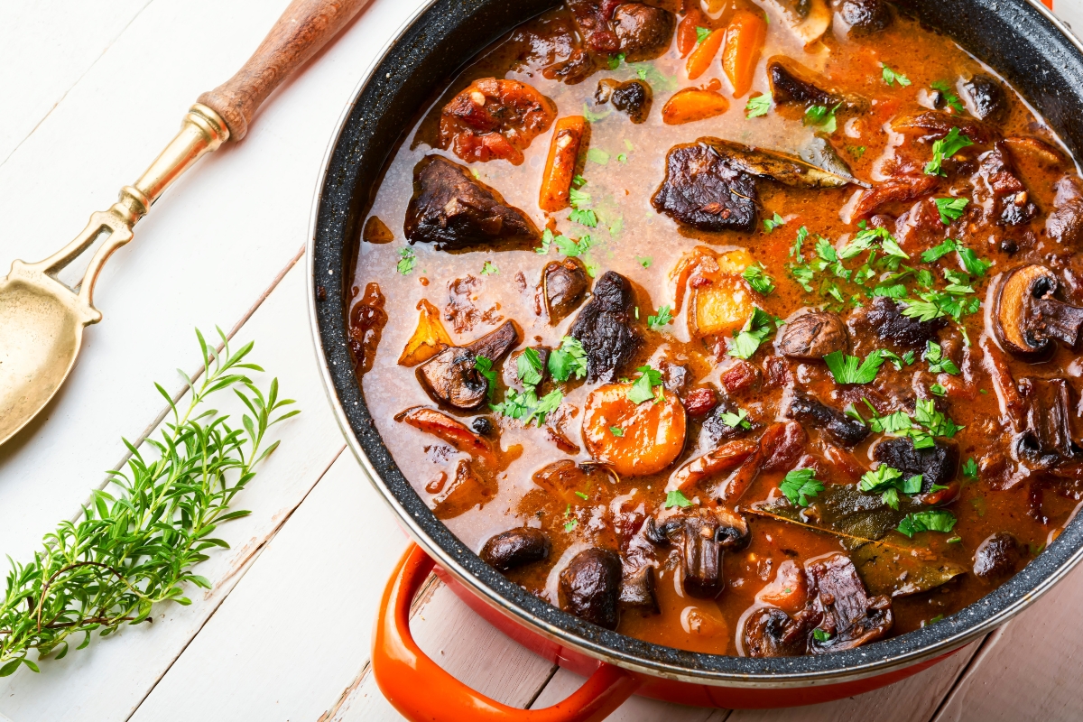 A red pot of hearty beef stew, brimming with carrots and mushrooms and garnished with parsley, sits atop a white wooden table. Perfect for a fall party food for a crowd, it rests beside a ladle and a sprig of rosemary.