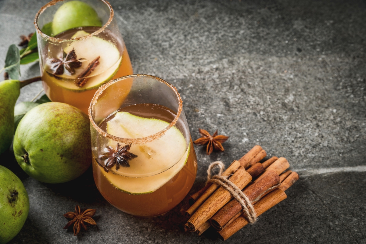 Two glasses of spiced apple cider with pear slices and star anise are perfect fall party food for a crowd. They sit invitingly beside fresh pears and tied cinnamon sticks on a gray surface, ready to delight guests with their warm flavors.