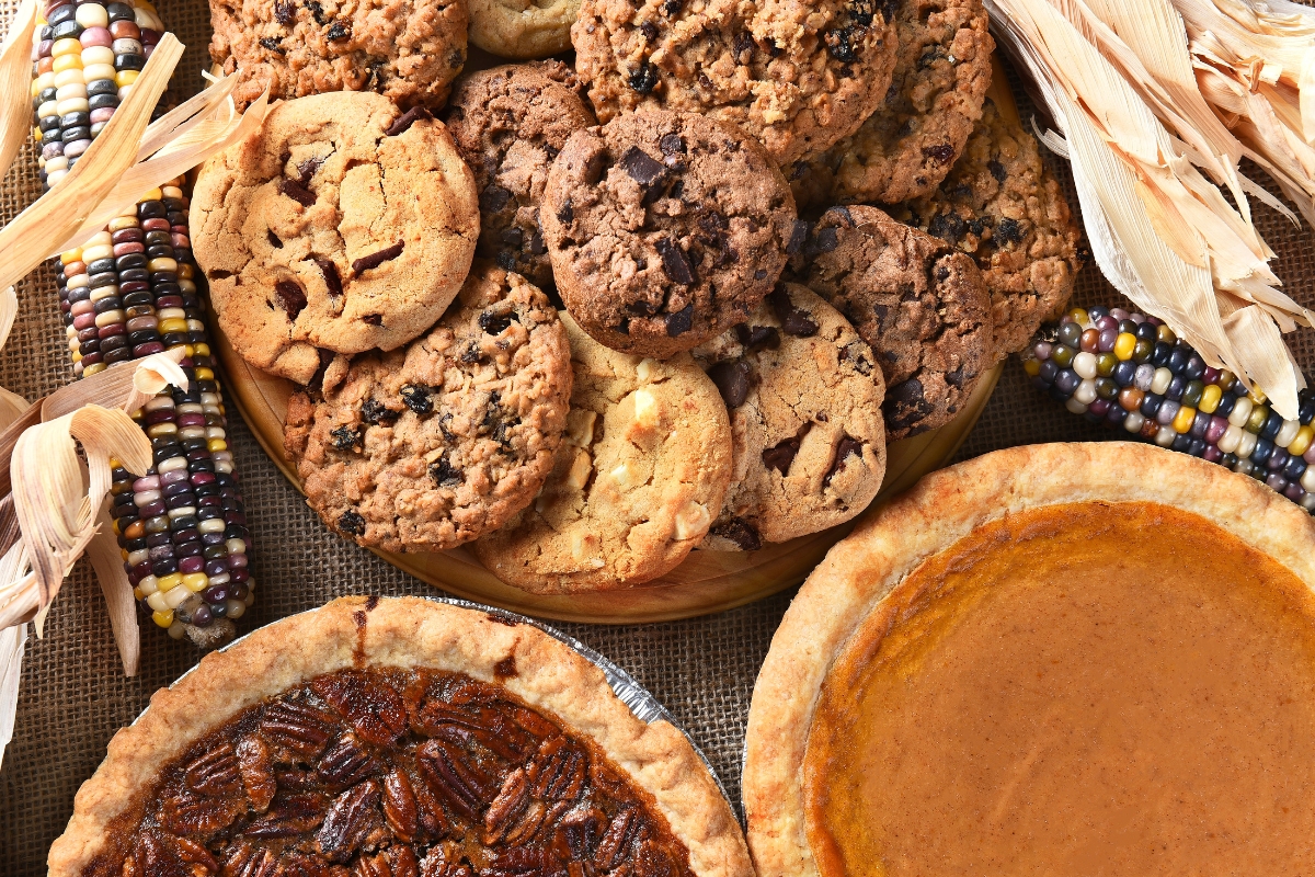 An assortment of cookies, a pecan pie, and a pumpkin pie on a table alongside decorative dried corn make the perfect fall party food for a crowd.