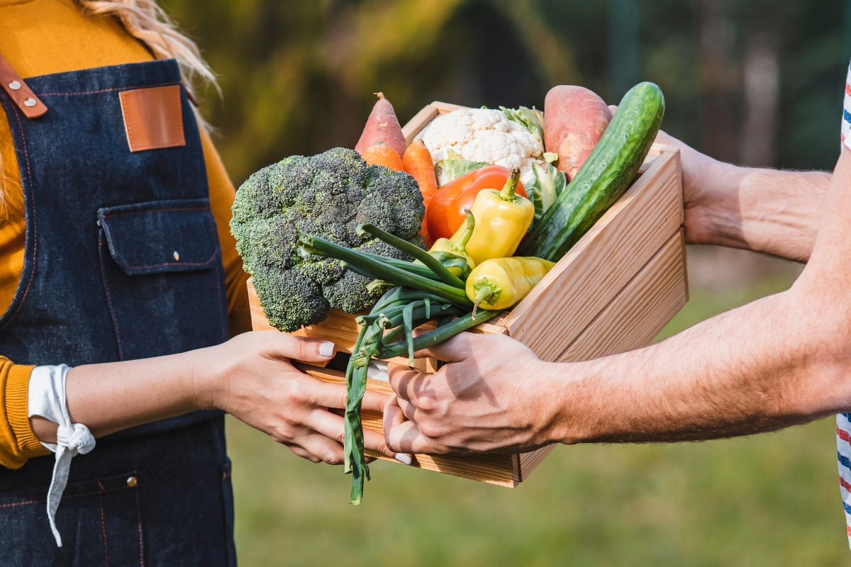 Two people exchanging a wooden box filled with fresh vegetables, including broccoli, peppers, cucumber, onions, and sweet potatoes—perfect for preparing fall party food for a crowd—outdoors.