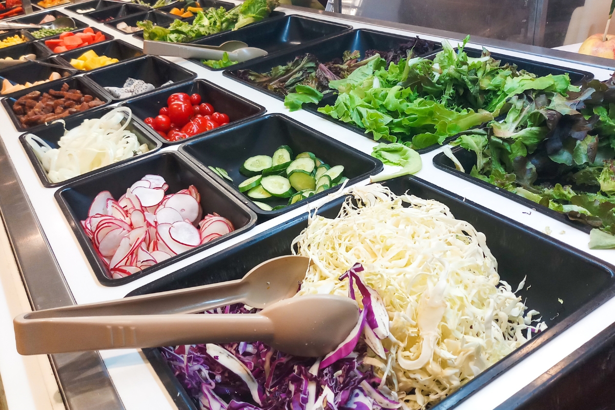 A salad bar perfect for a fall party, featuring fresh ingredients like lettuce, cucumber, radishes, cherry tomatoes, sliced onions, shredded cabbage, and bell peppers—all beautifully displayed in black containers with tongs to serve a crowd.