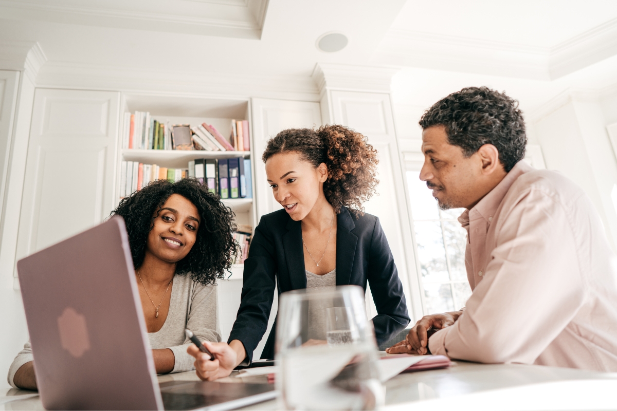 Three people are gathered around a laptop in an office setting, collaborating on event planning. One person is pointing at the screen while discussing catering options, and the others are watching and listening attentively.