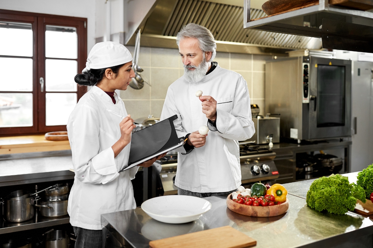 Two chefs in a kitchen, one holding a clipboard and pen, planning an upcoming event's catering menu, while the other holds a garlic bulb. A bowl of fresh vegetables sits invitingly on the counter.