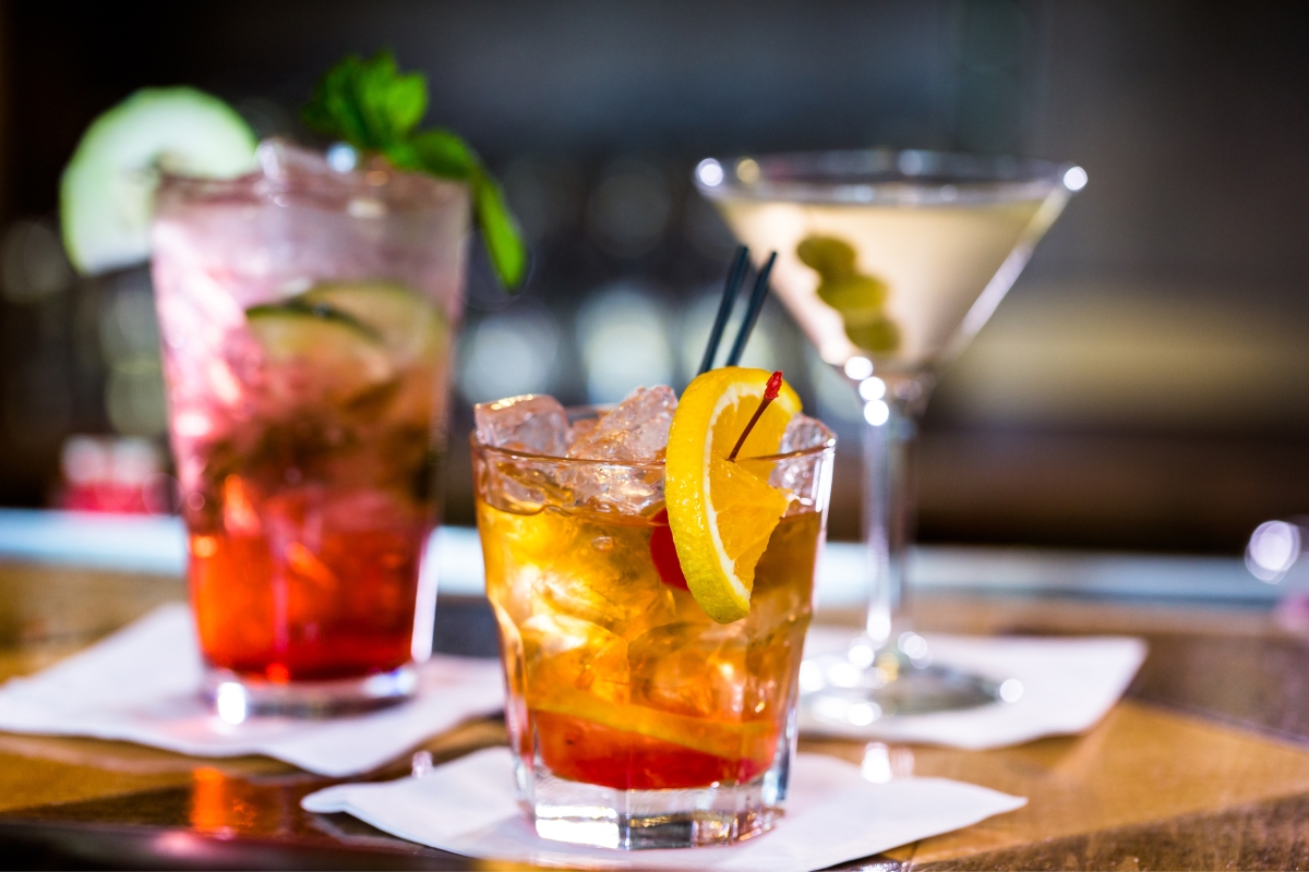 Three cocktails elegantly displayed on a bar: a red beverage with cucumber and mint, an amber delight garnished with a lemon slice and straw, and a clear martini adorned with olives. Perfect for event planning and catering, these drinks add sophistication to any gathering.