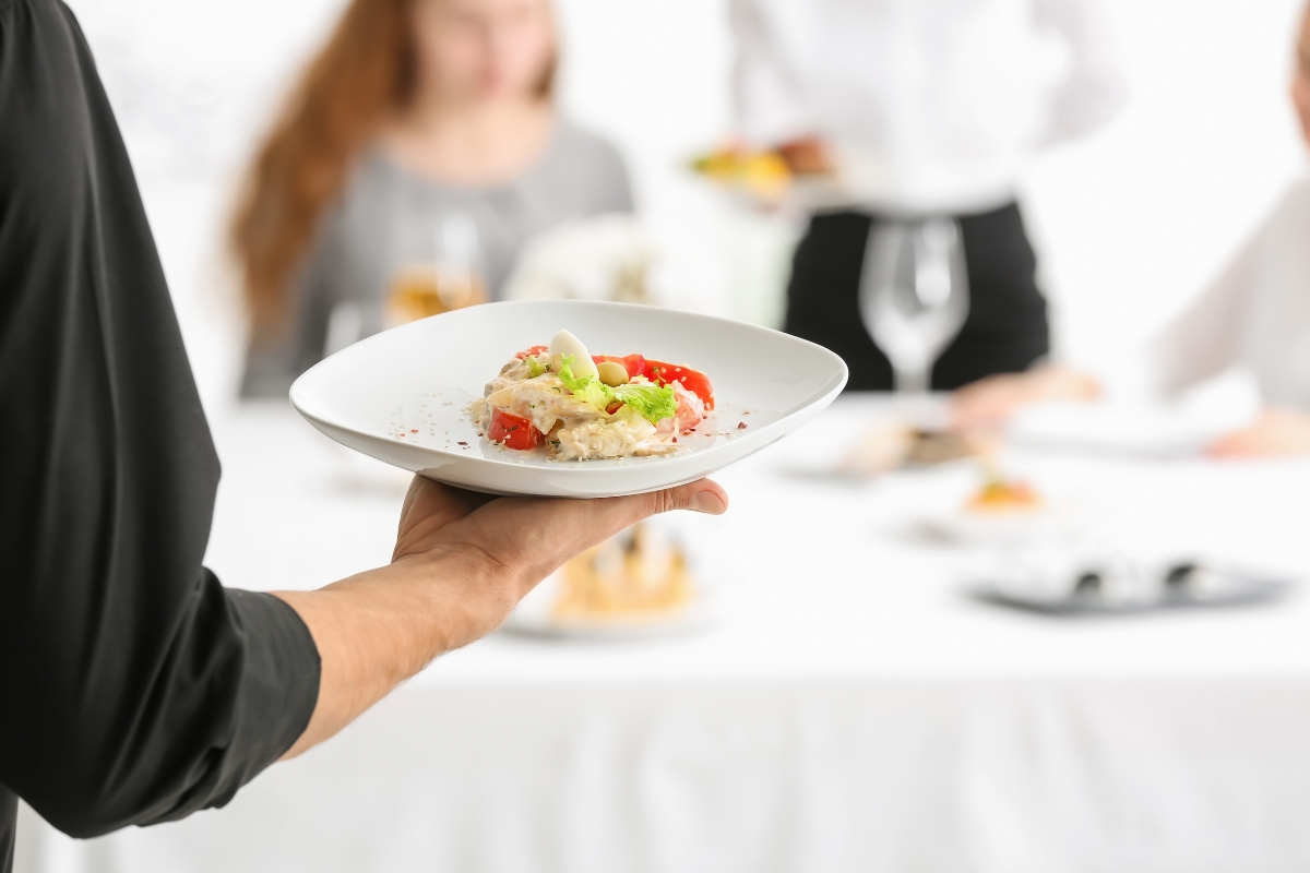 A person, skilled in event planning, is elegantly serving a plate of risotto garnished with tomatoes and herbs at a dining table, where blurred figures in the background savor the culinary expertise.