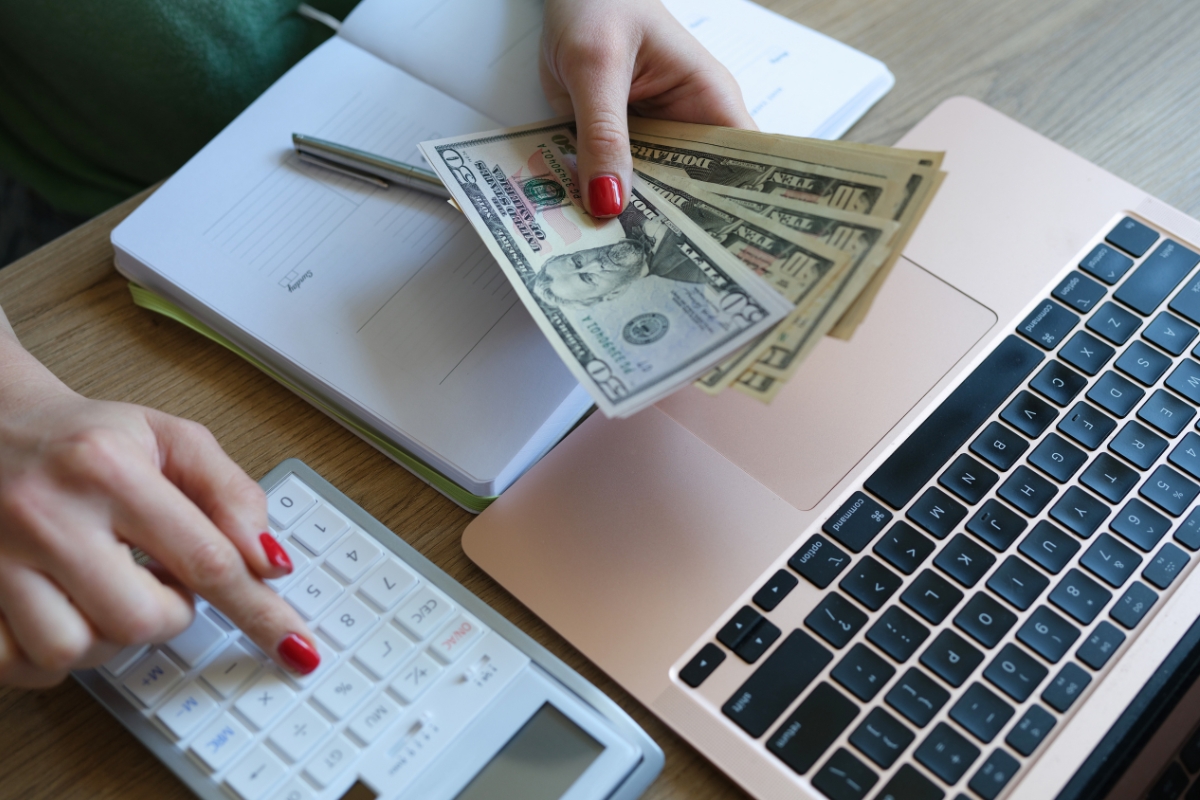 A person, amidst event planning and catering tasks, holds US dollar bills while meticulously using a calculator and laptop on a wooden desk.