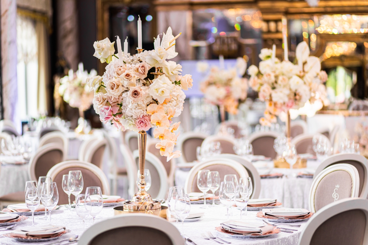 Elegant wedding reception with tables set for dining, featuring white and blush floral centerpieces, surrounded by white chairs in a lavishly decorated room. Expert event planning ensures seamless coordination while catering delights guests with an exquisite culinary experience.