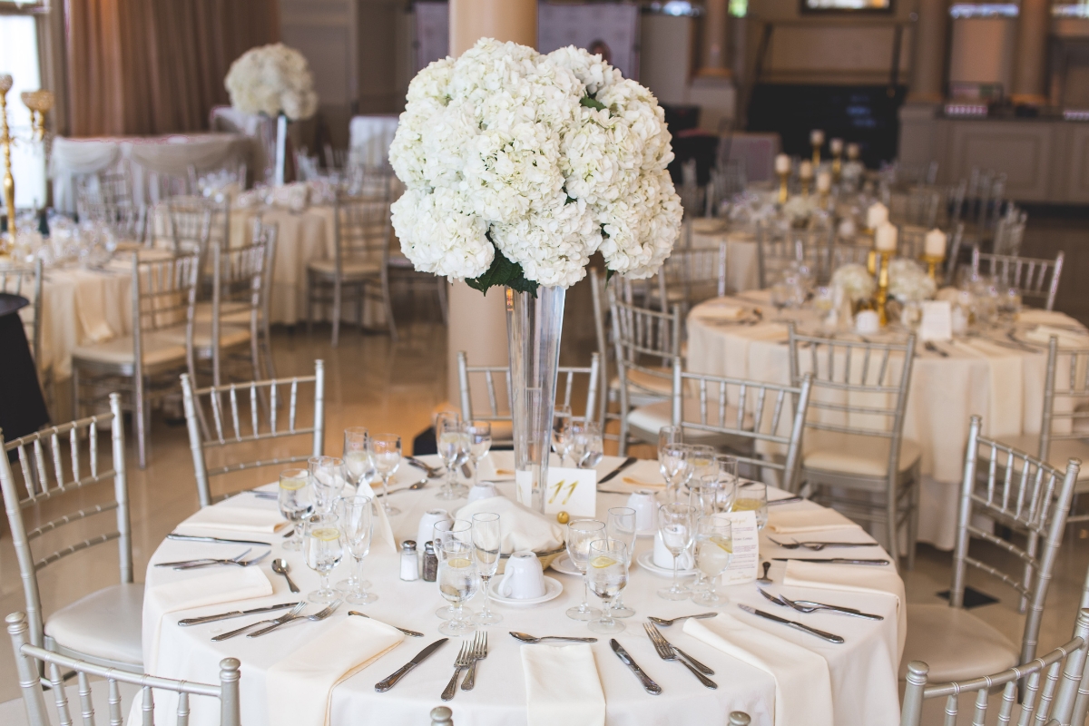 Catering Blog 1 A dining table set for an elegant event showcases a wedding catering menu, with a white floral centerpiece, silver chairs, and meticulously arranged place settings in a beautifully decorated venue.