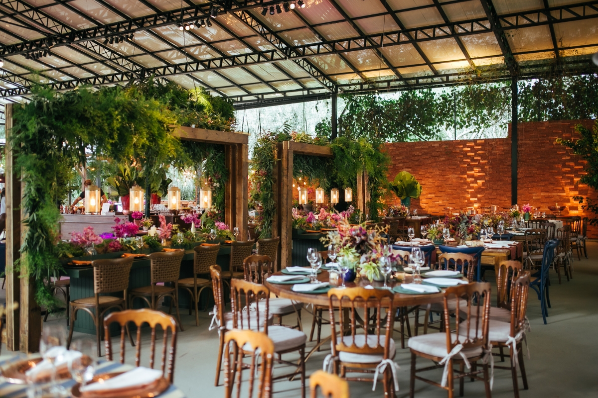 Rustic indoor event space with limited venue availability, featuring wooden tables, chairs, and greenery decor. Tables set with plates, glasses, and floral centerpieces under a glass roof. Warm lighting throughout.