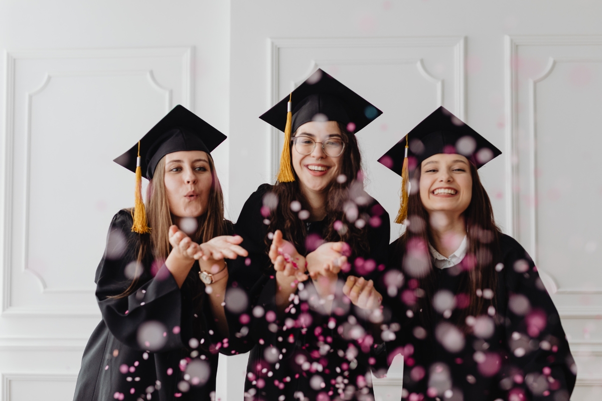 Three graduates in caps and gowns smile and blow pink confetti towards the camera against a white background, celebrating their achievements—perfect inspiration for those planning catering for graduation parties.