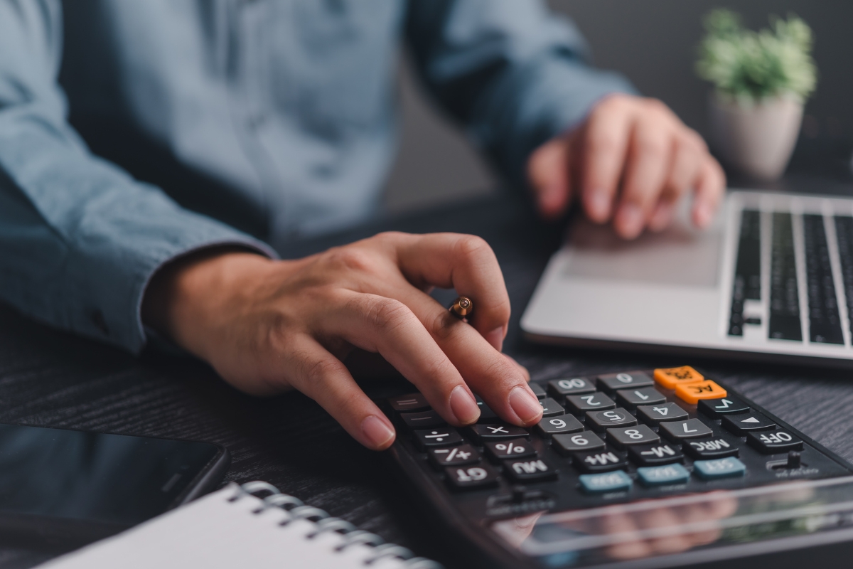 Catering for Graduation Parties Made Easy: Stress-Free Planning Tips 9 A person uses a calculator with one hand and a laptop with the other, planning catering for graduation parties next to a notepad and smartphone on a desk.