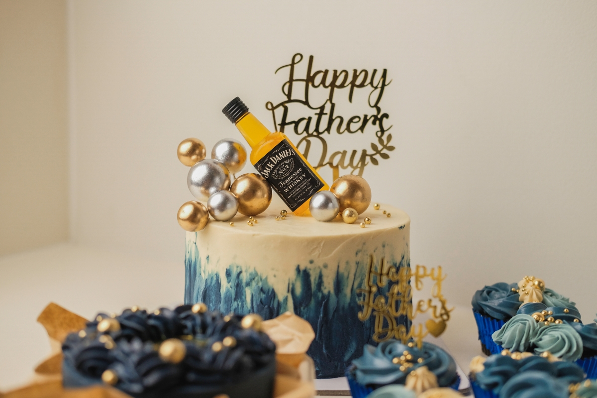 A decorated cake with a "Happy Father's Day" topper, gold and silver spheres, and a small Jack Daniel's bottle, perfect for Father's Day catering, surrounded by blue and gold cupcakes and treats.