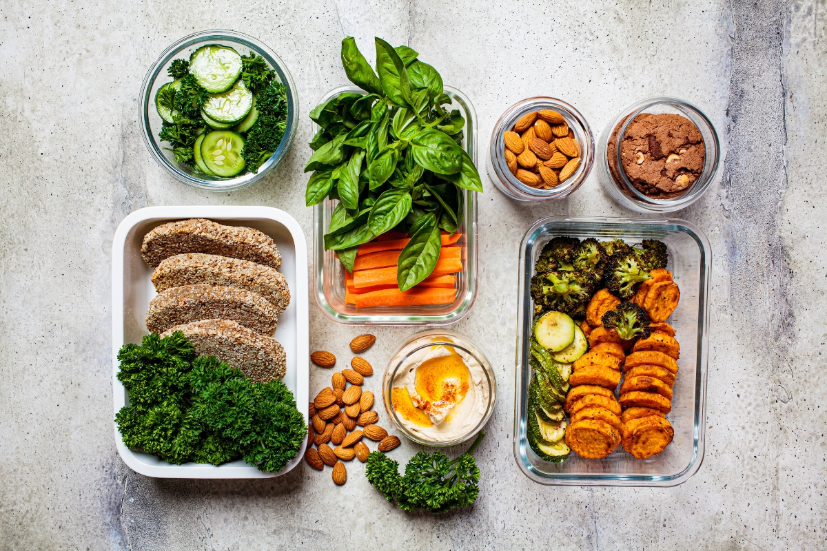 Father's Day Catering: Menu Ideas to Honor Dad 7 Assorted healthy foods including sliced bread, leafy greens, carrot sticks, basil, hummus, almonds, chocolate powder, cucumber, and roasted vegetables arranged in glass containers—perfect for Father's Day Catering.