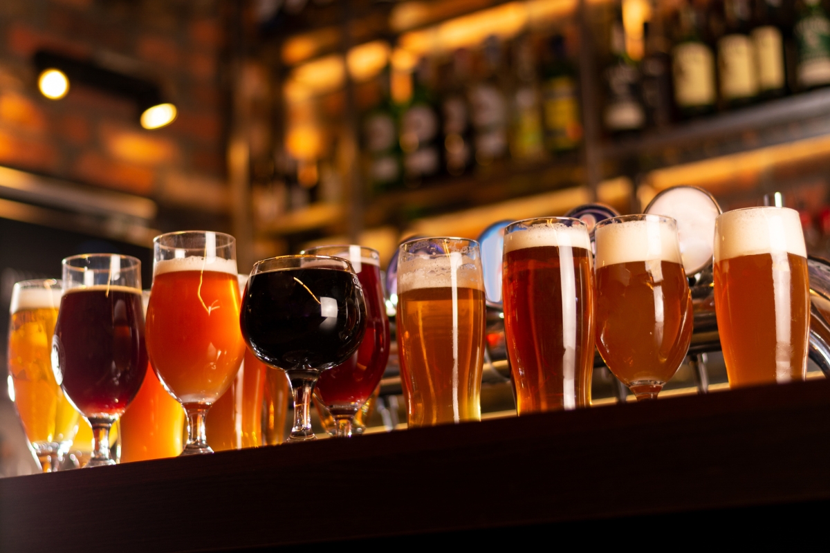 Father's Day Catering: Menu Ideas to Honor Dad 8 A row of assorted beer glasses filled with different types of beer sits on a bar counter, perfect for Father's Day catering, with shelves of bottles blurred in the background.