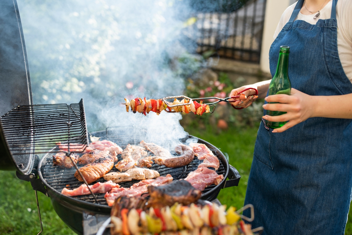 Father's Day Catering: Menu Ideas to Honor Dad 1 A person in a denim apron grills assorted meats and vegetable skewers on a charcoal barbecue while holding a green bottle, perfect for Father's Day Catering.