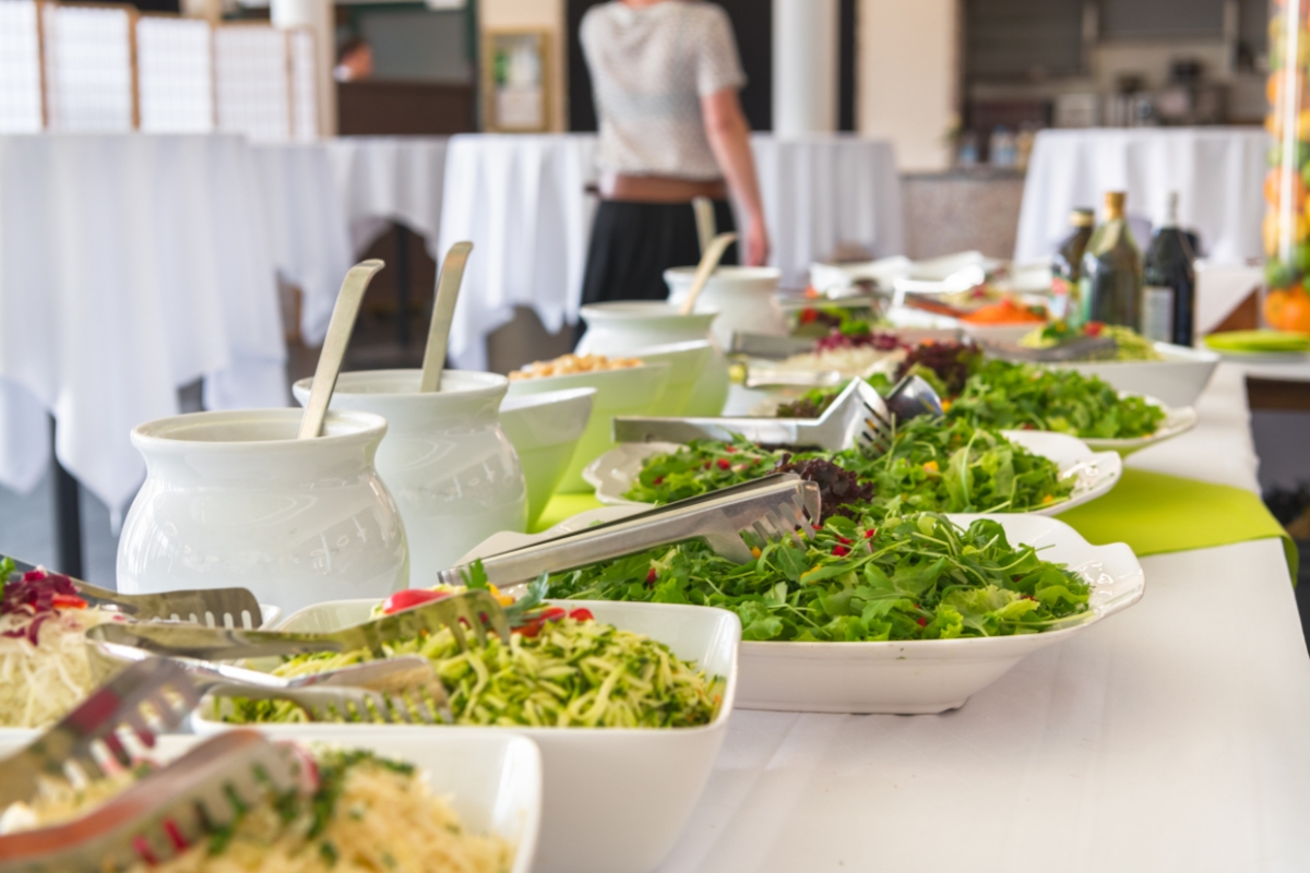 Healthy Catering Options: Light and Fresh Menus for Summer Events 5 A buffet table with various salads, greens, and salad dressings in jars offers healthy catering options in a bright indoor space with white tablecloths.