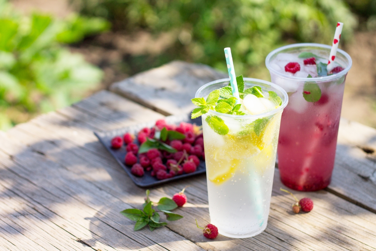 Healthy Catering Options: Light and Fresh Menus for Summer Events 6 Two plastic cups with fruit drinks—one lemon and mint, the other with raspberries and mint—sit on a wooden table beside a plate of fresh raspberries outdoors, showcasing healthy catering options.