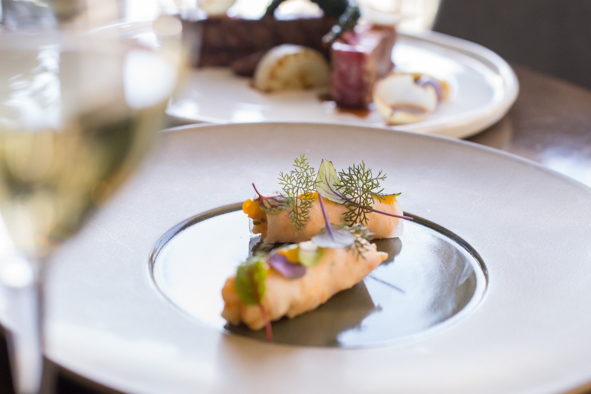 Healthy Catering Options: Light and Fresh Menus for Summer Events 8 A gourmet dish with delicately plated seafood rolls garnished with herbs, set on a white plate—an elegant example of healthy catering options—with another dish and a glass of white wine in the background.