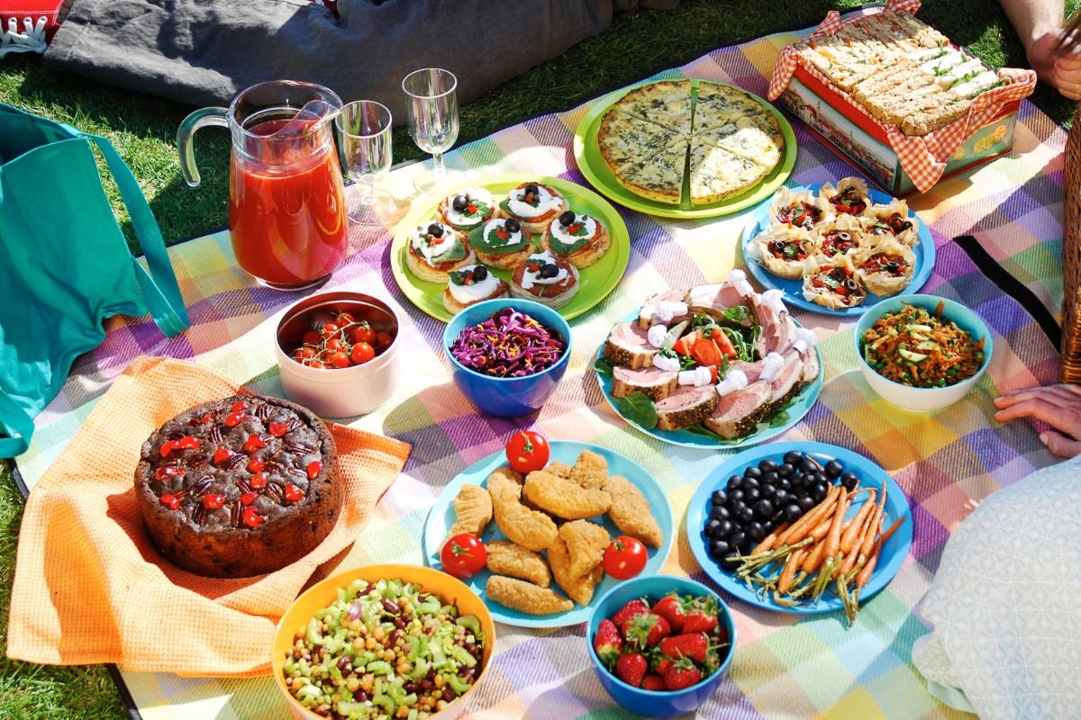Patriotic Catering: Fourth of July Menu Ideas to Impress 3 A colorful picnic spread on a blanket showcases assorted dishes, perfect for Fourth of July Menu Ideas—salads, cake, tart, bread, a pitcher of drink, and bowls of fruit and vegetables create a festive feast.