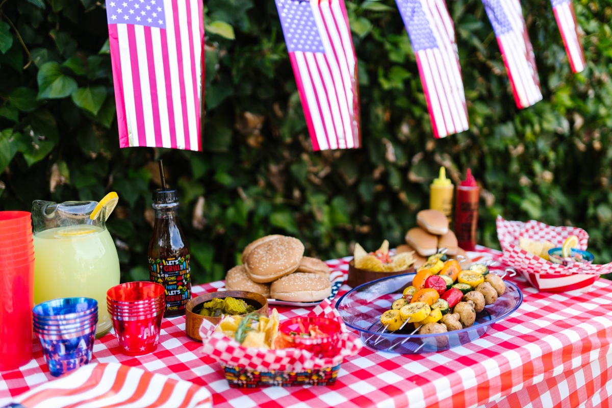 Patriotic Catering: Fourth of July Menu Ideas to Impress 5 A picnic table with red checkered tablecloth is set with burgers, skewers, drinks, condiments, and cups, decorated with small American flags hanging above—perfect for gathering Fourth of July Menu Ideas.