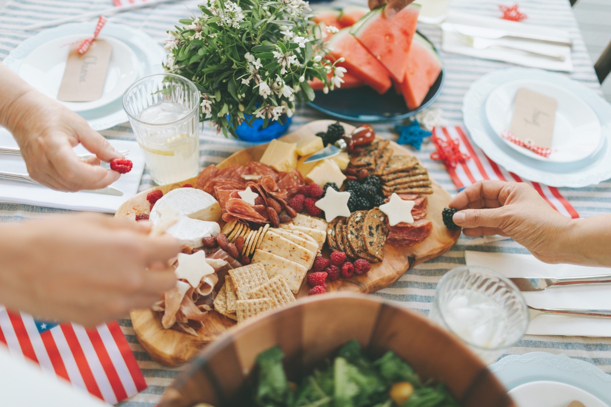 Patriotic Catering: Fourth of July Menu Ideas to Impress 7 A table set with a charcuterie board of crackers, cheese, fruit, and meats—perfect for Fourth of July menu ideas—flanked by salad, watermelon slices, drinks, and utensils. Hands are reaching for food.
