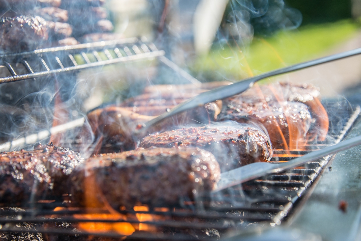 Patriotic Catering: Fourth of July Menu Ideas to Impress 1 Close-up of hamburger patties cooking on a grill with visible smoke; metal tongs are turning one of the patties—perfect inspiration for your Fourth of July menu ideas.