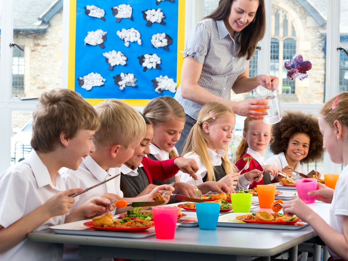 Back to School Dinner Ideas for Educational Events 6 A teacher pours water for a group of children eating lunch at a school cafeteria table, with colorful cups and plates of food in front of them—perfect inspiration for back to school dinner ideas.