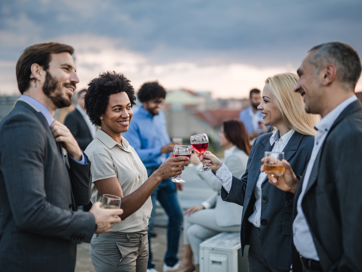 A group of professionally dressed adults stand outdoors, enjoying drinks and smiling while clinking glasses during a gathering with Corporate Picnic Catering.