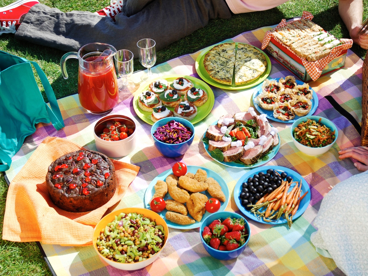 A picnic spread on a blanket features salad, cake, bread, fruit, chicken nuggets, tomato dishes, drinks, and various side dishes with people sitting around—perfect for memorable Corporate Picnic Catering events.