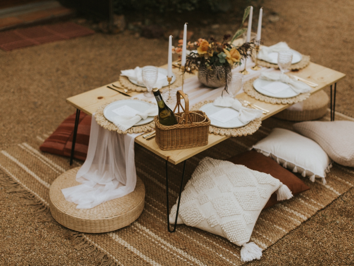 A low wooden picnic table set with plates, glasses, candles, and flowers offers a charming setting for corporate picnic catering, surrounded by floor cushions and pillows on a woven rug outdoors.
