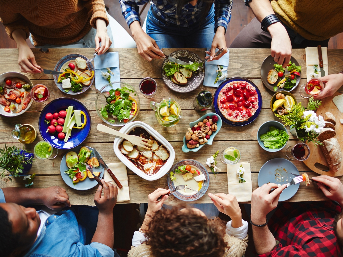 A group of people enjoying a meal together at a wooden table set with various dishes, salads, drinks, and desserts—perfect for a Corporate Picnic Catering event.