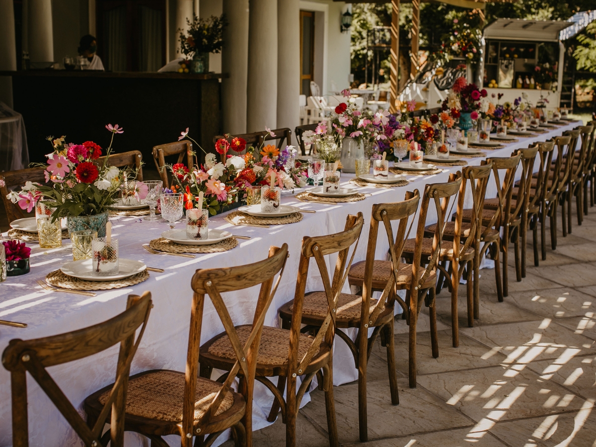 A long banquet table with wooden chairs is set outdoors, decorated with colorful flower arrangements, glassware, and plates—perfectly prepared for a formal event or upscale Corporate Picnic Catering experience.